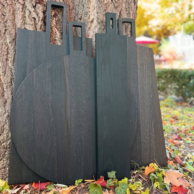 
                  
                    Shot of a collection of blackened oak serving boards leaning against a tree outdoors, in autumn. 
                  
                